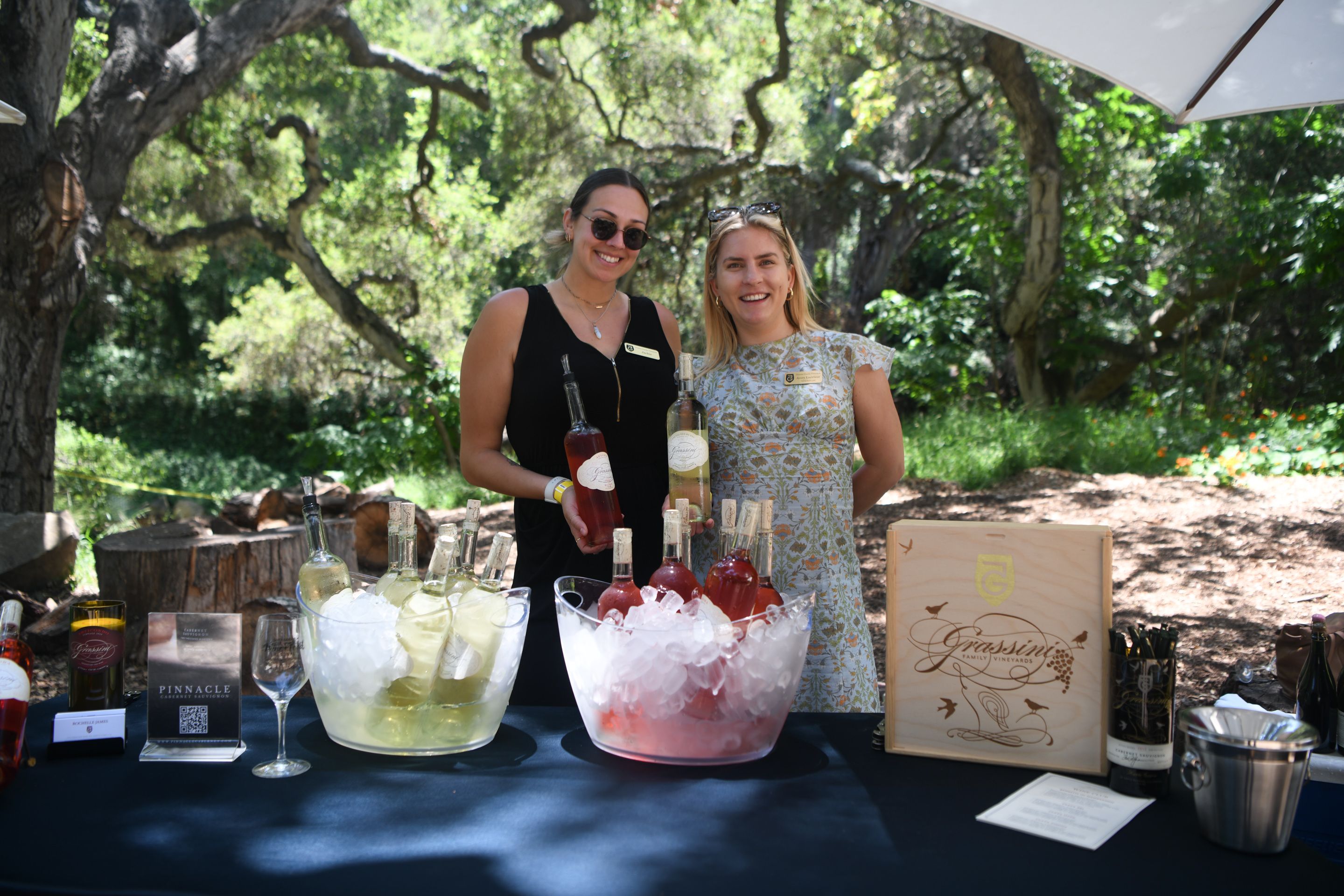 Two women surrounded by trees holding winer bottles at a wine festival.